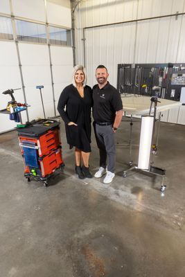 Woman and man smiling in a shop with tools; woman wears black dress, man a black polo, and concrete floor.