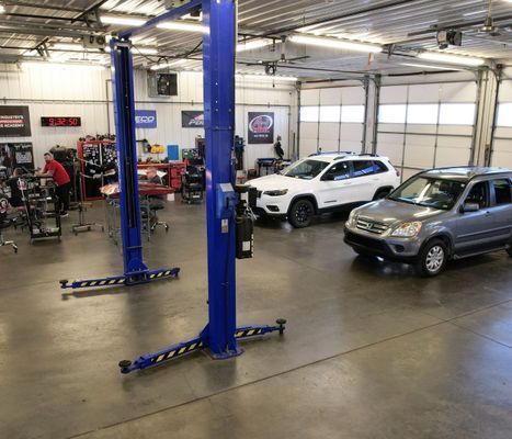 Inside a car repair shop, two SUVs sit, near a lift. A worker is in the background.