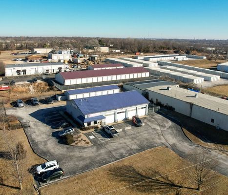 Aerial view of a storage facility with multiple buildings, a blue-roofed office, and parked cars on a sunny day.