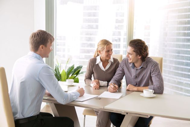 Three people in a bright office meeting, talking around a table with papers and coffee cups.
