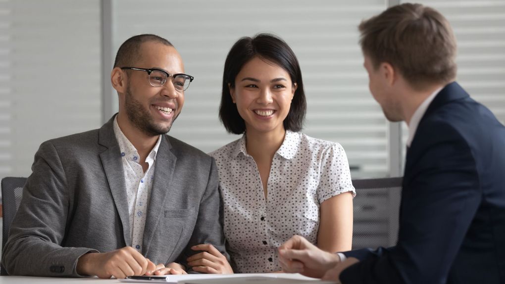 Three coworkers in a meeting, smiling and talking at a table in an office.