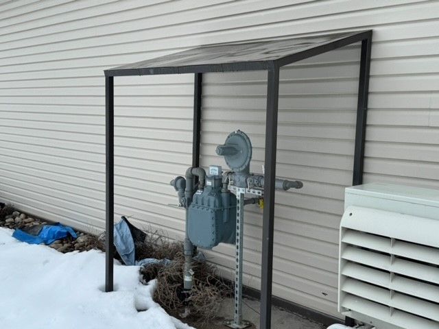 A gas meter is sitting under a canopy on the side of a house.