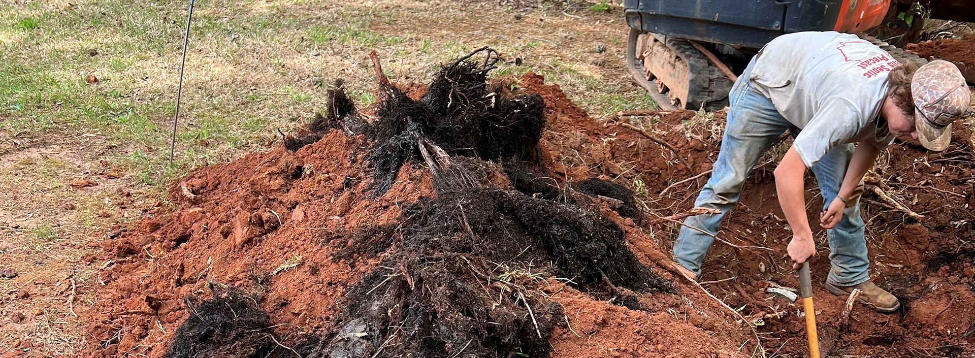 Person digging a mound of dark material with a shovel beside a trailer outdoors