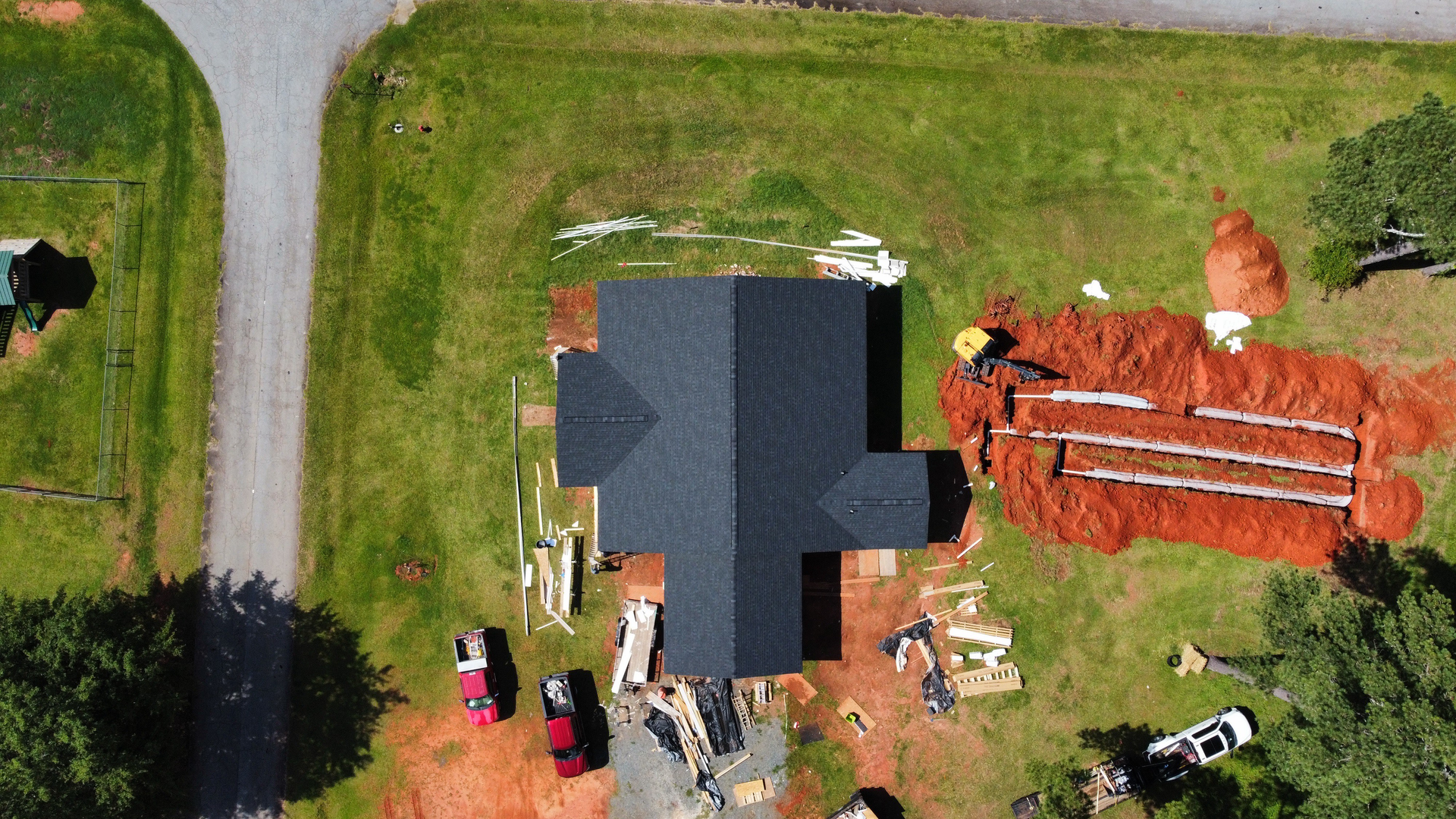 Aerial view of a house under construction with a dark roof, surrounded by materials, vehicles, and red dirt.