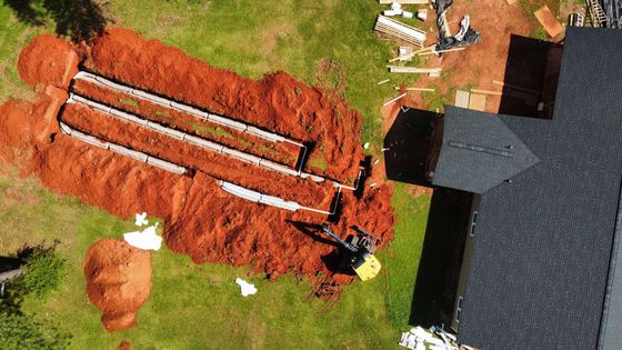 Learn more about drain lines Aerial view of a construction site with trenches dug in red earth, likely for underground utilities, next to a house with a dark roof.