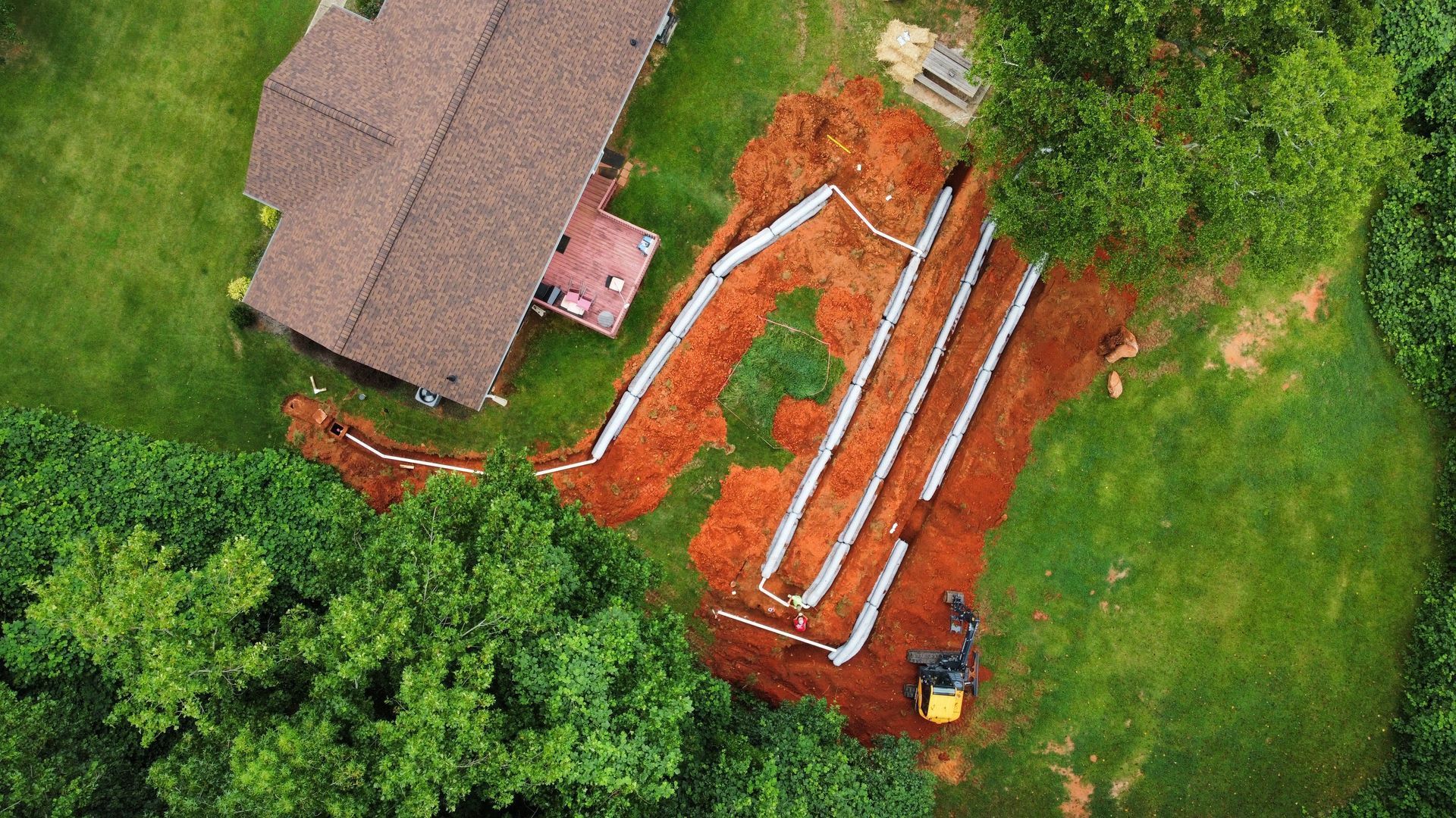 Overhead view of a house with a drainage system being installed. Trenches filled with white pipes snake across the red-brown earth.