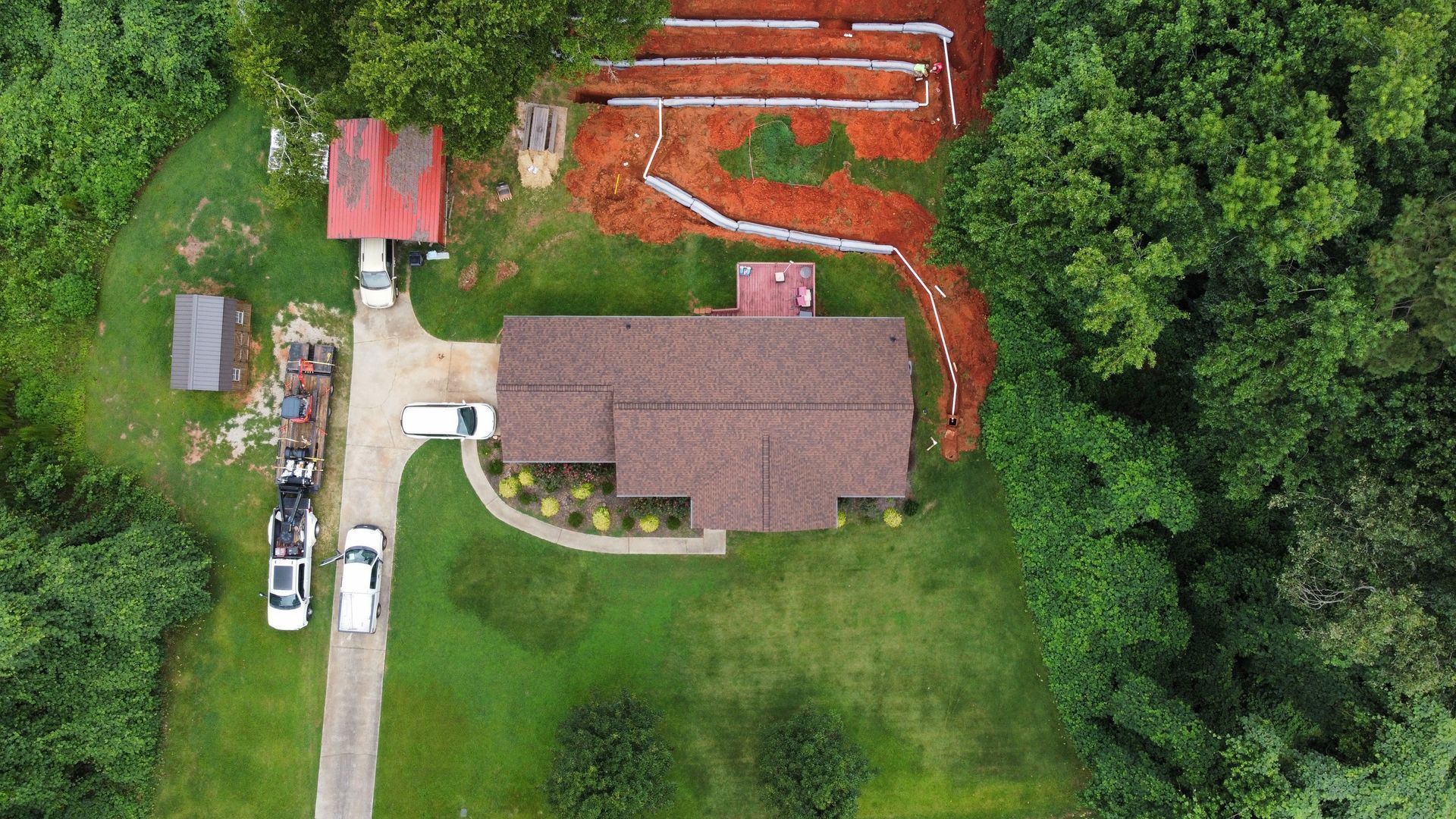 Aerial view of a house with a brown roof and surrounding landscaping, with a driveway and vehicles. Trees and a red shed are nearby.