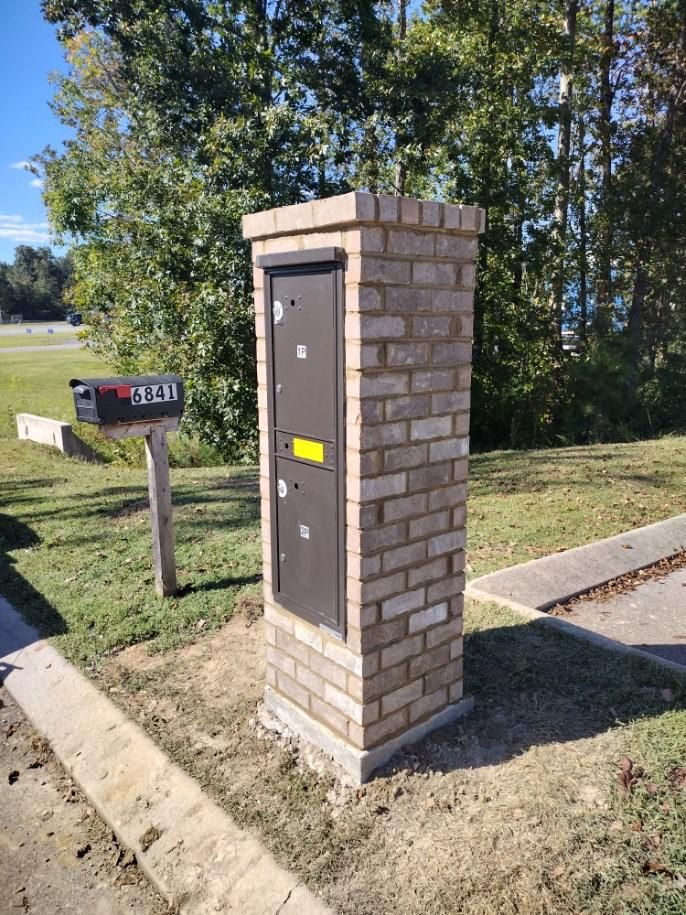 A brick mailbox pillar next to a curb and a grassy area, with a black mailbox in the background.