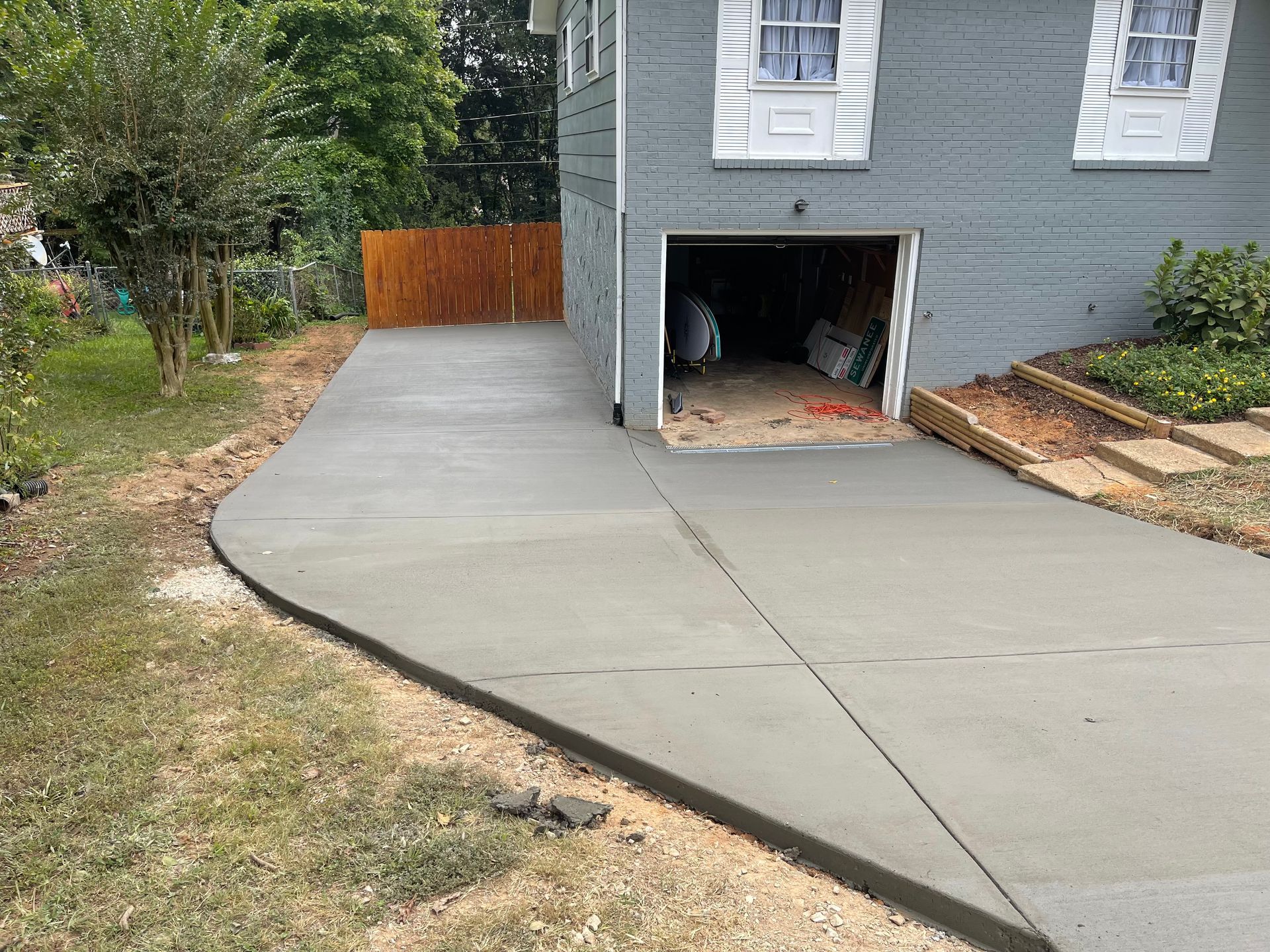 Newly poured concrete driveway leading to a gray house with an open garage, with a wooden fence in the background.