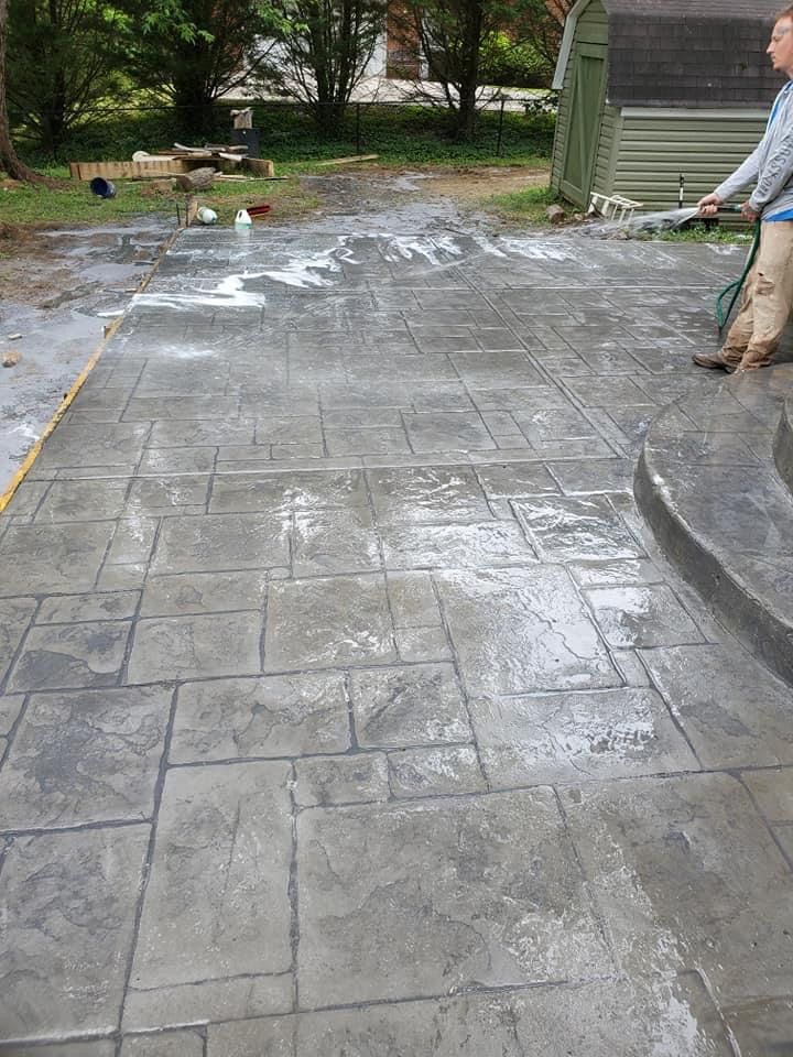 Person washing newly laid, stamped concrete patio with a hose; gray, brick-patterned surface.
