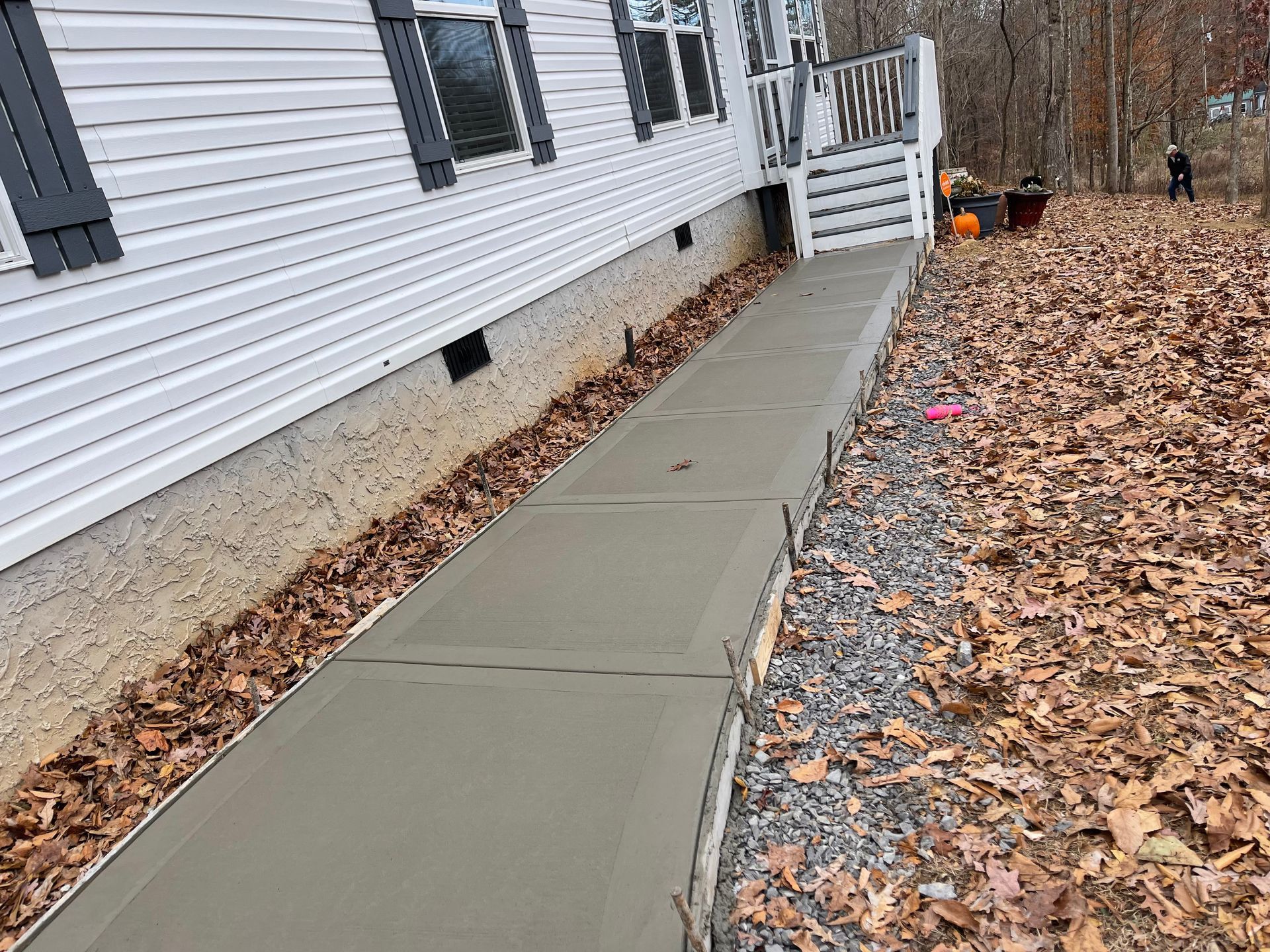 Newly poured concrete walkway next to a white house with grey shutters, with a person in the distance.