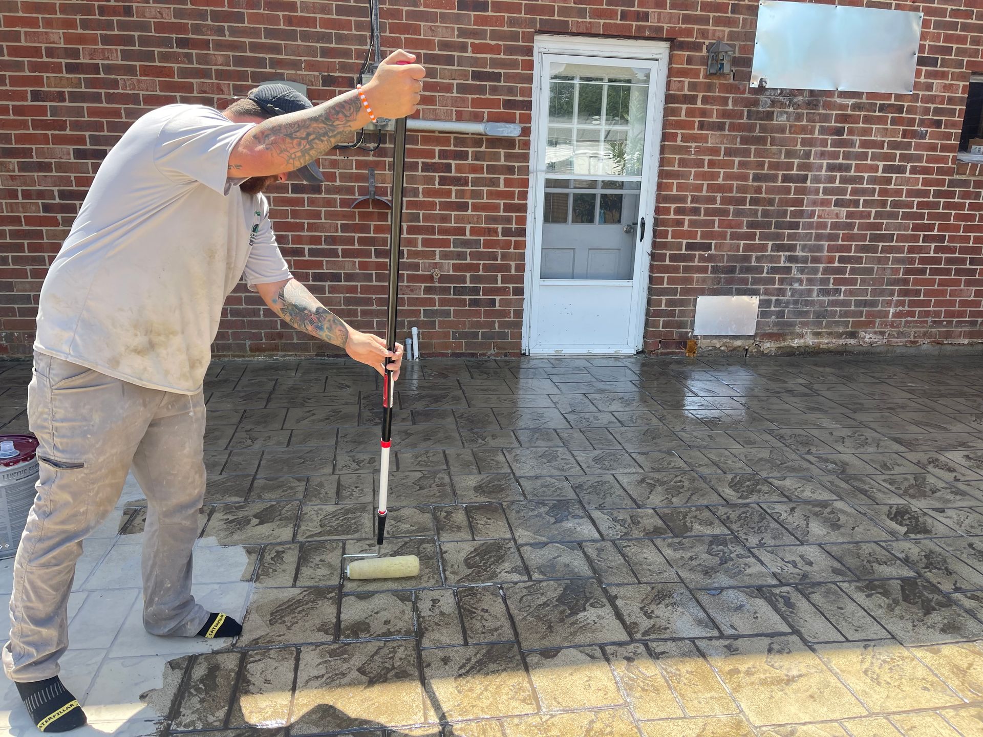 A person rolling sealant on stamped concrete patio near a brick building.