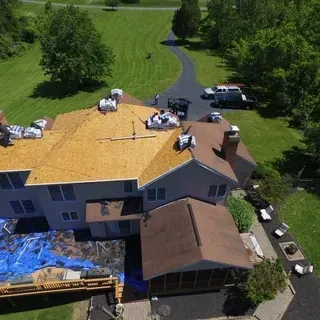 An aerial view of a house undergoing roof renovations, showing exposed wooden decking and stacks of new roofing shingles.