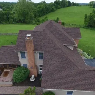 Aerial view of a house with a dark brown shingled roof, a brick chimney, and surrounding green lawn.