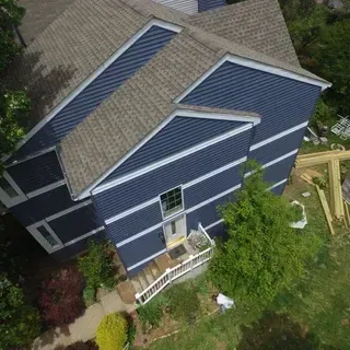 An aerial view of a blue, two-story house with a grey shingled roof, a front porch, and a yard with construction materials.