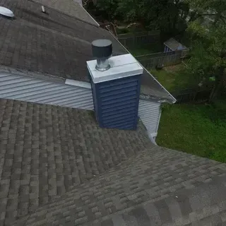 A high-angle view of a gray asphalt shingle roof featuring a blue, square-sided chimney chase with a metal top and cap.