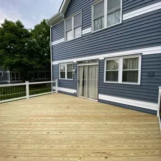 A large, newly built light wood deck attached to the back of a blue-sided house with white trim and windows.