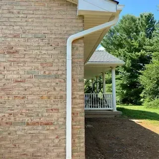 A vertical white downspout mounted against a tan brick wall next to the porch of a house.