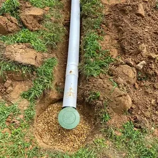 A grey plastic drainage pipe ending in a circular green cap, set in a dirt trench filled with gravel.