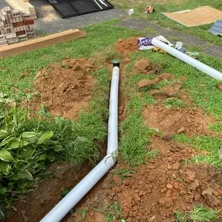 A trench in a grass yard contains white PVC drainage pipe with a green cleanout cap, with dirt piles alongside.