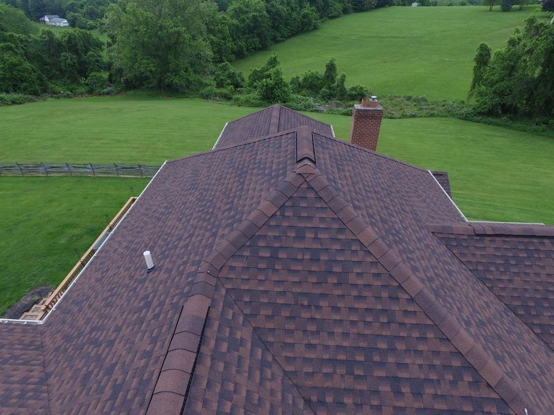 An aerial view of a brown shingled roof on a house, surrounded by a green field and trees.