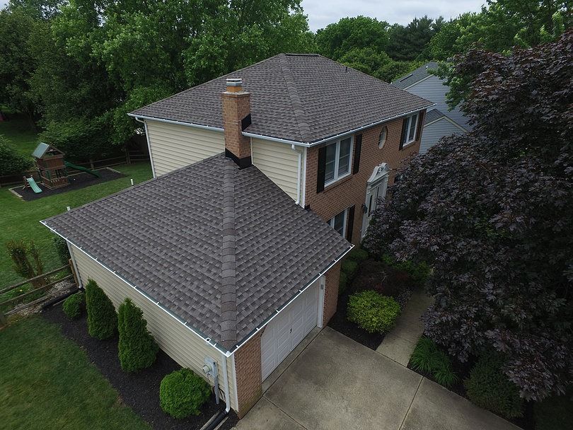 Aerial view of a two-story home with a brick and beige facade, dark roof shingles, and a surrounding lawn with trees.
