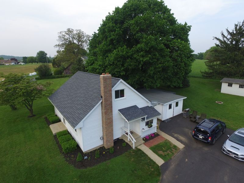 An aerial view of a white, one-story house with a grey roof, brick chimney, large tree, and driveway with two parked cars.