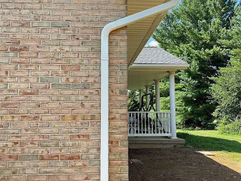A tan brick wall corner with a white downspout, next to a porch with white columns and a railing near trees.
