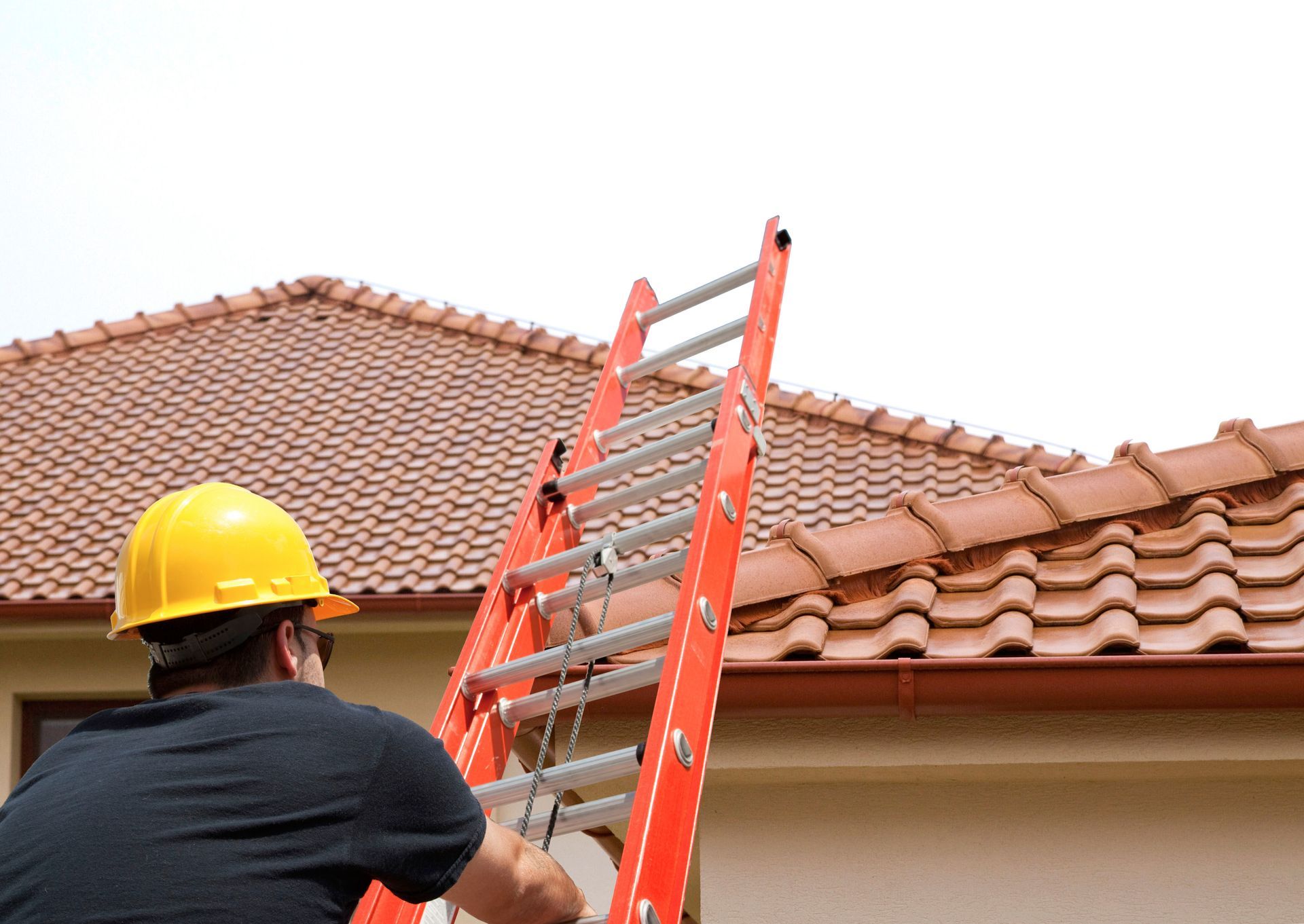 Person in yellow hard hat on a ladder, ascending towards a tiled roof.