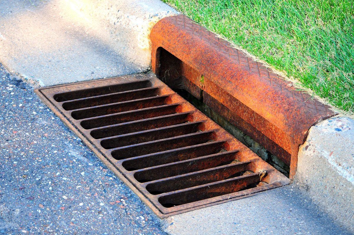 Rusty metal storm drain at the edge of a sidewalk next to grass.