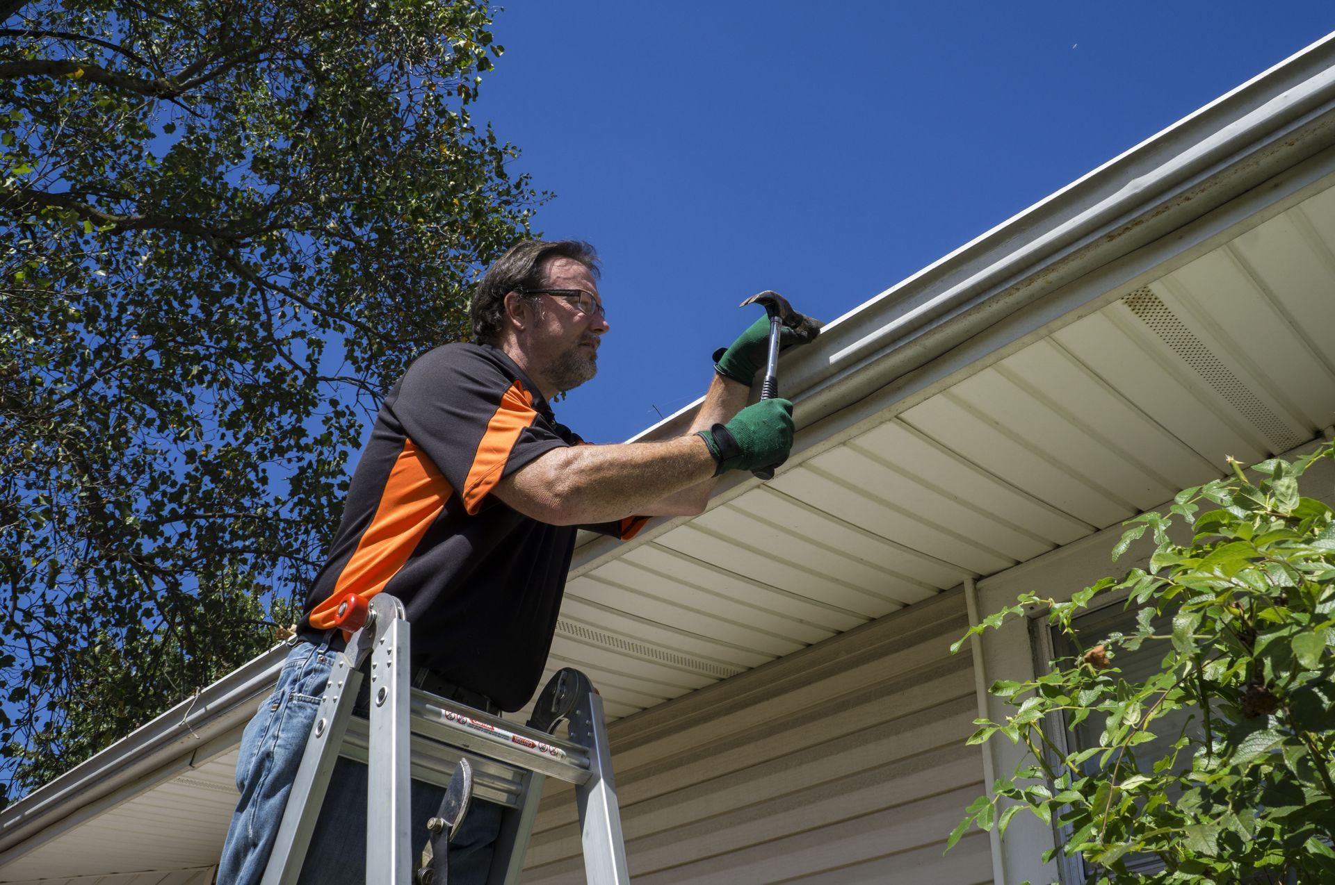 Man on a ladder cleaning a gutter with a screwdriver. Blue sky and foliage in the background.
