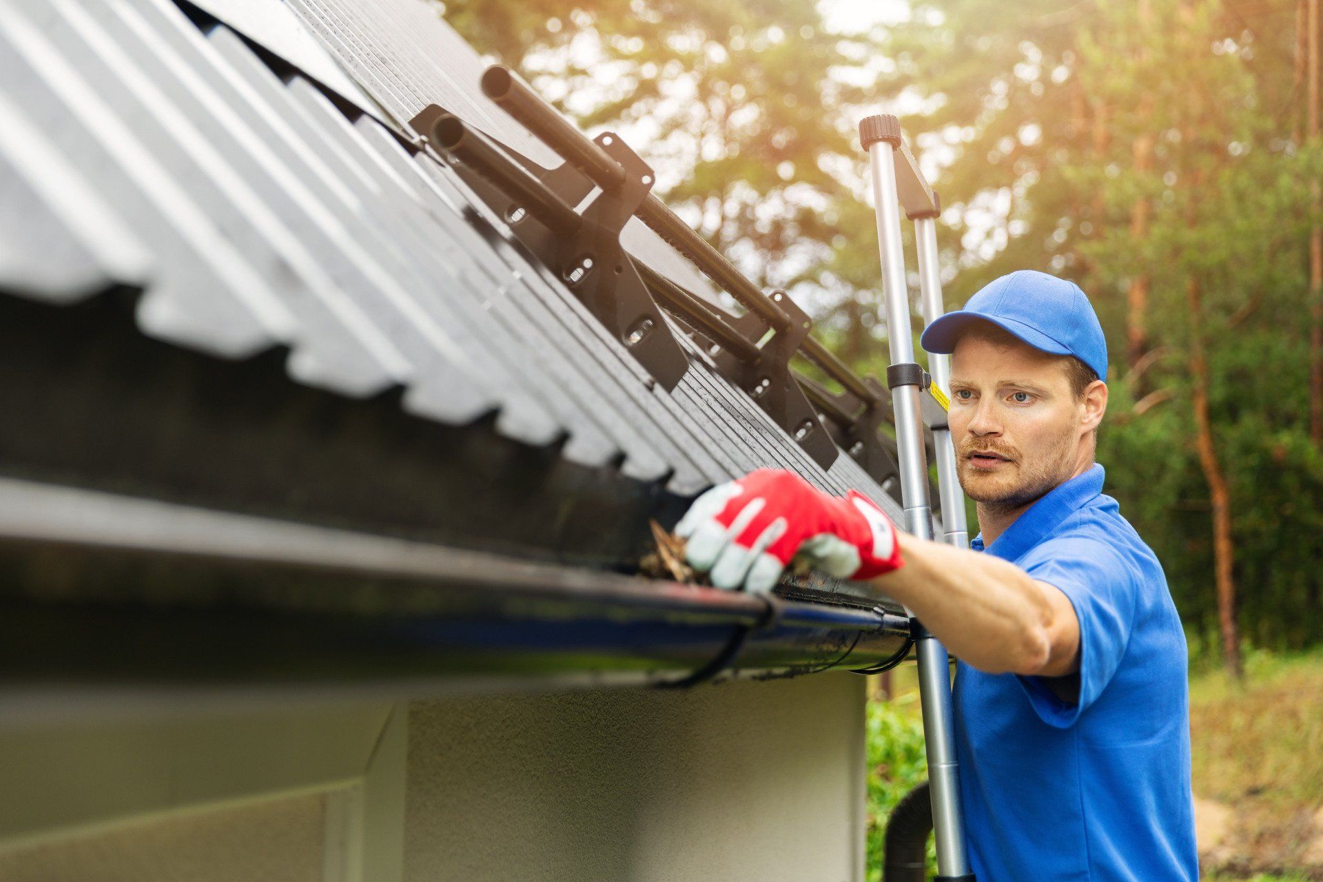 Man in blue shirt and cap cleaning gutters with a ladder outdoors.