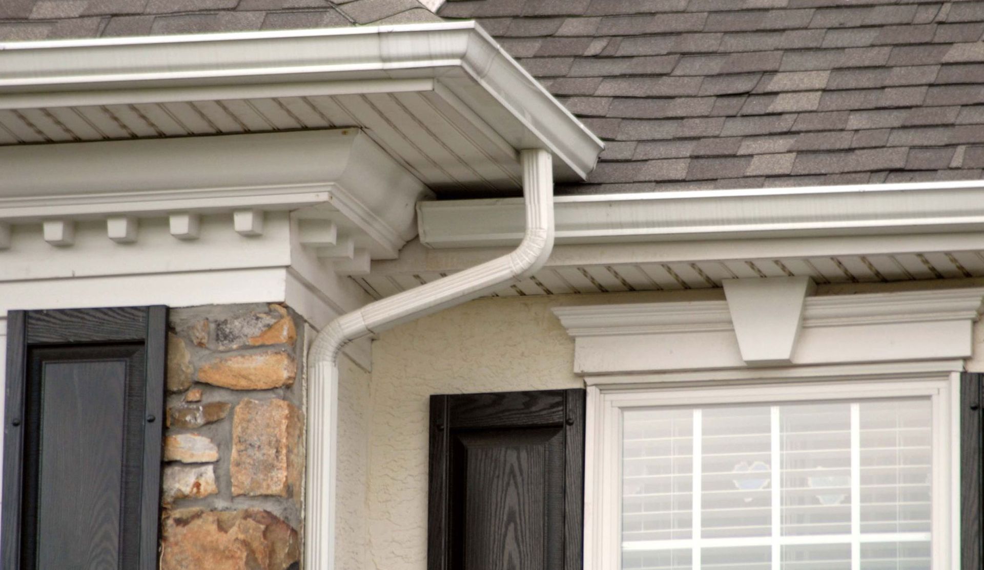 White gutters and downspout on a house with a stone facade, shutters, and a shingled roof.