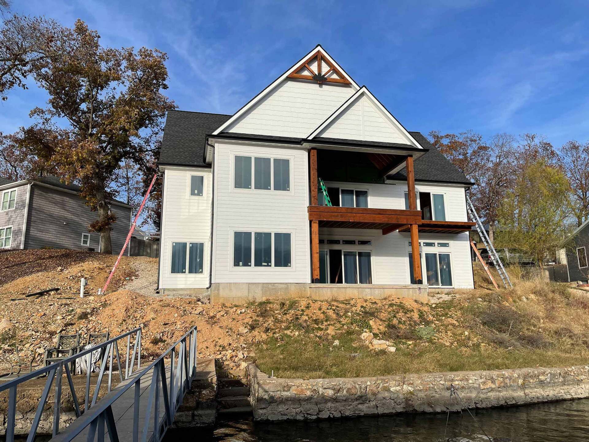 Two-story house under construction on a hillside near water; white siding, brown trim, and a dark roof.