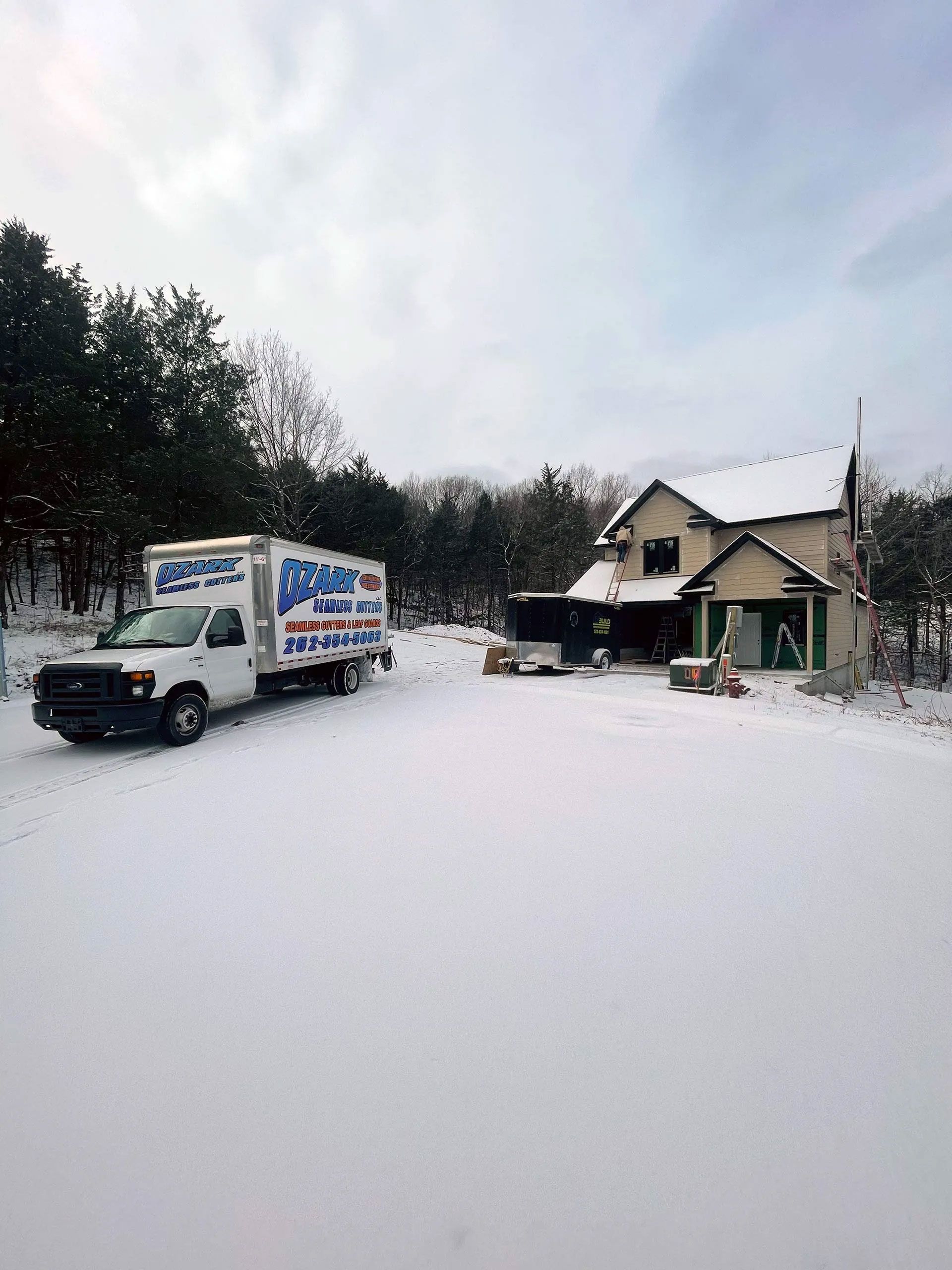 White moving truck parked in snowy yard near a two-story house, surrounded by trees under a cloudy sky.