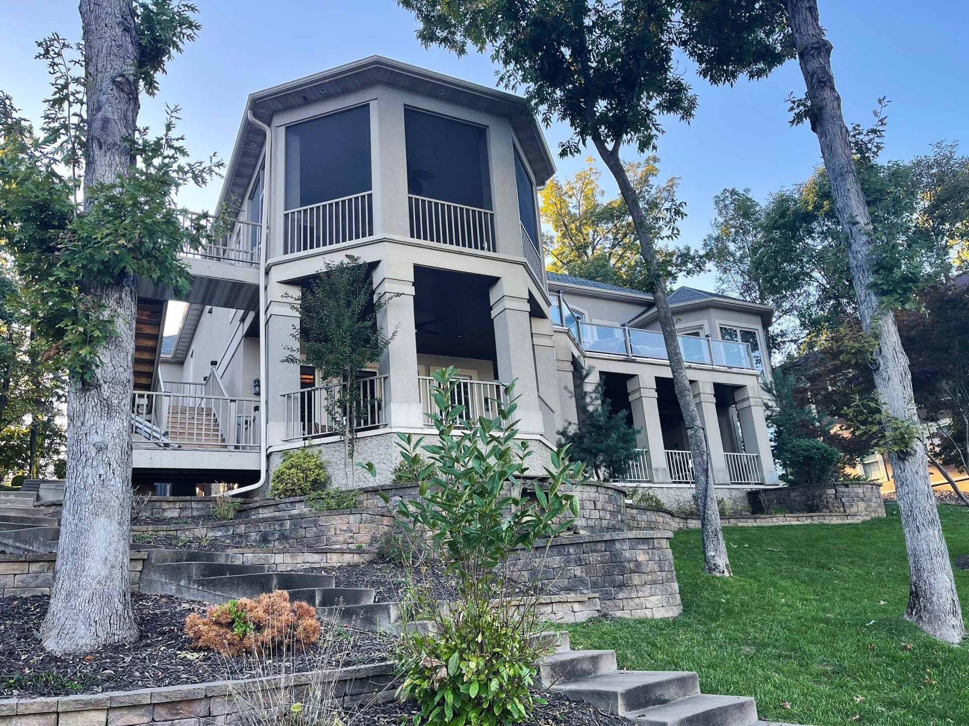 Two-story house with columns and balconies, set on a grassy hill near trees and a stone wall.
