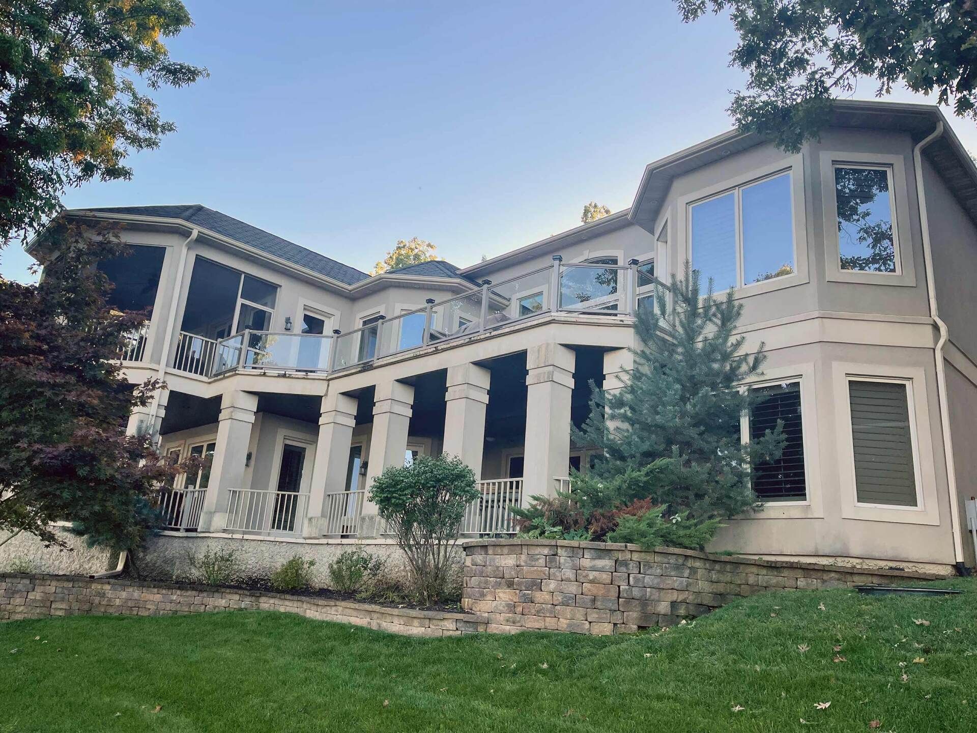 Two-story beige house with large windows, columns, and a lawn. Trees and sky are in the background.