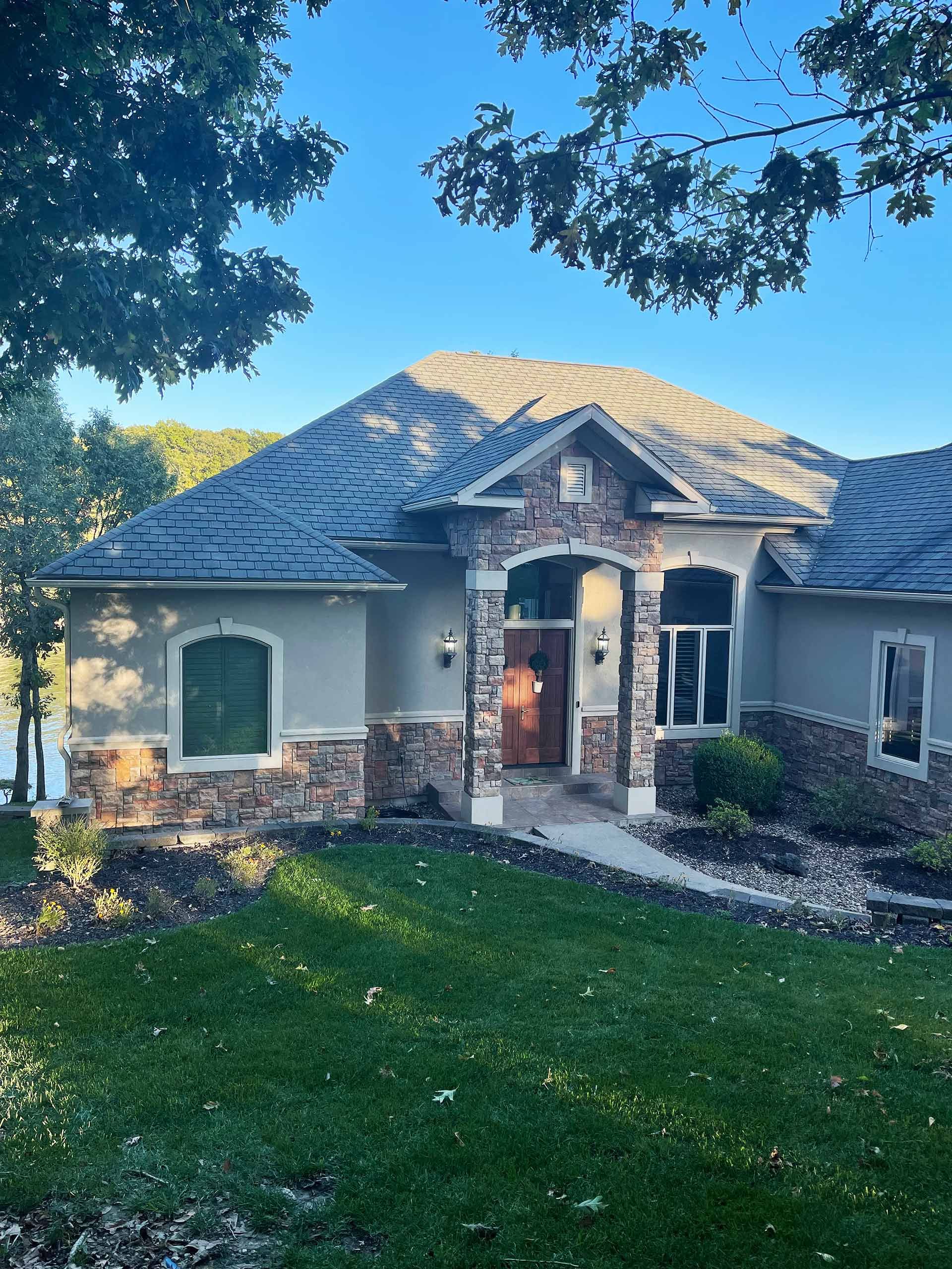 Stone-accented, beige stucco house with a dark gray roof. Green lawn and trees in the foreground. Blue sky.