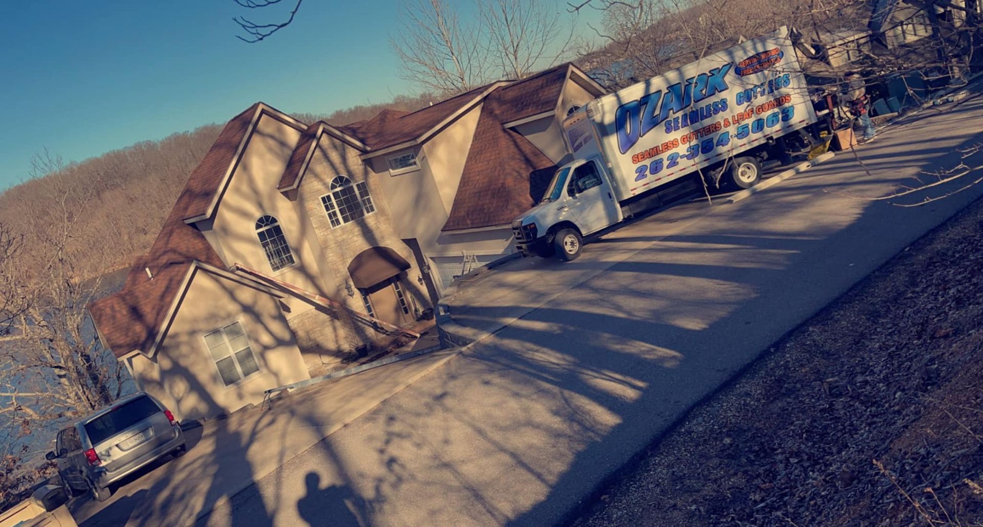 A house and a moving truck parked on a sloped road, in a sunny setting with shadows.