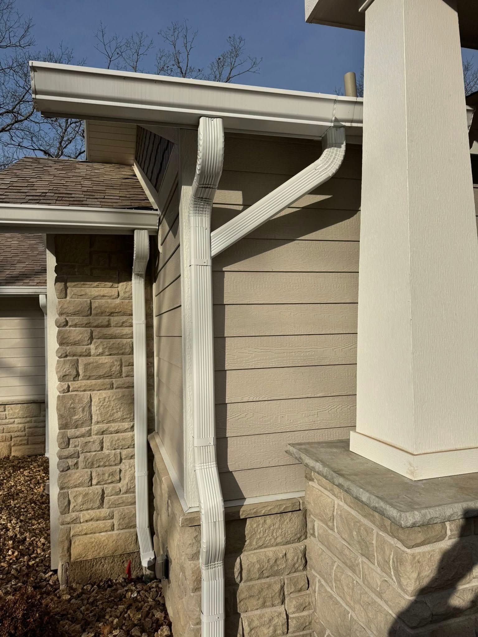 White gutter and downspout on a light-colored house with stone and siding, beneath a clear, blue sky.