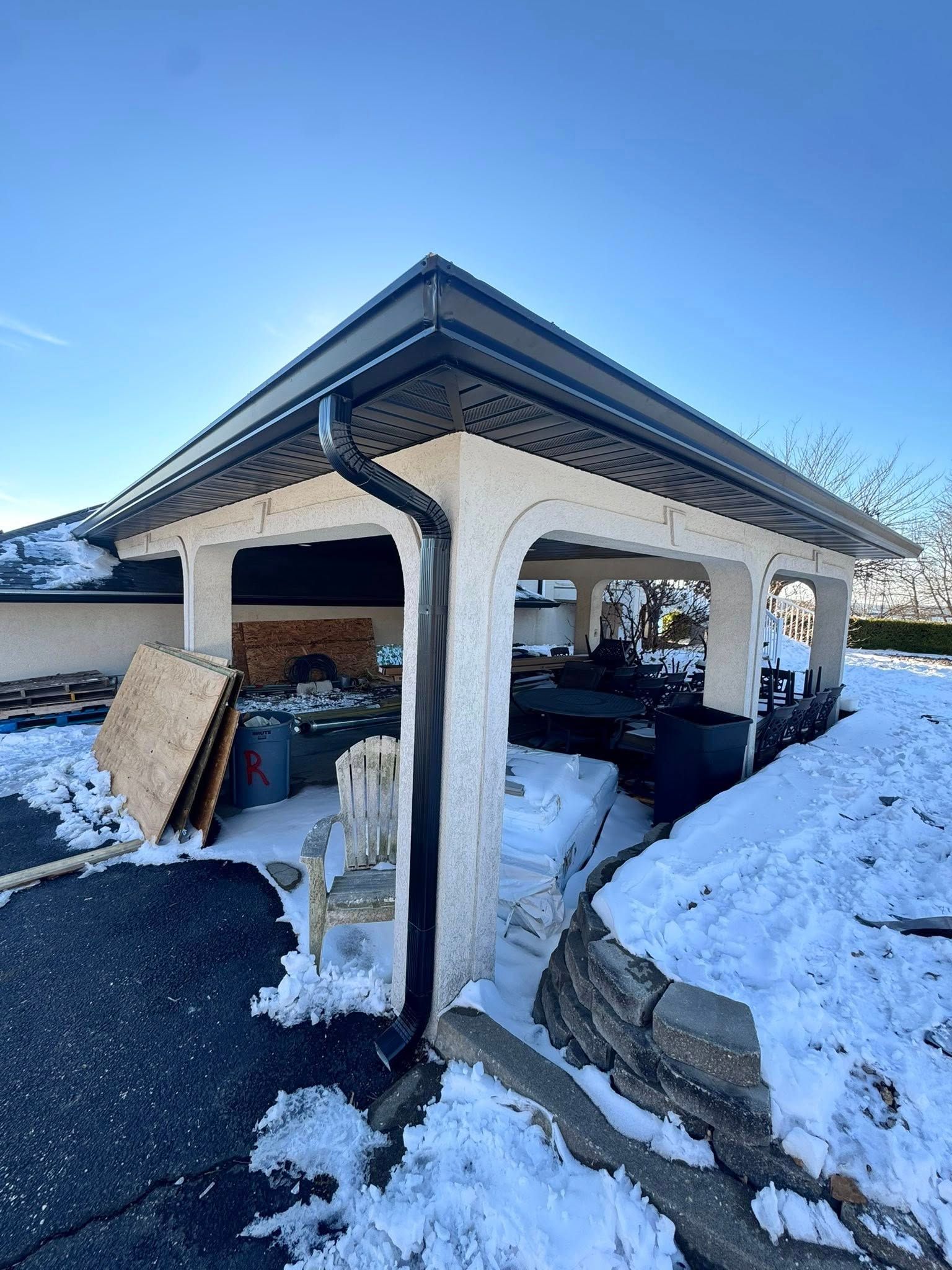 Covered outdoor structure with dark roof and snow on the ground; plywood and other materials nearby.