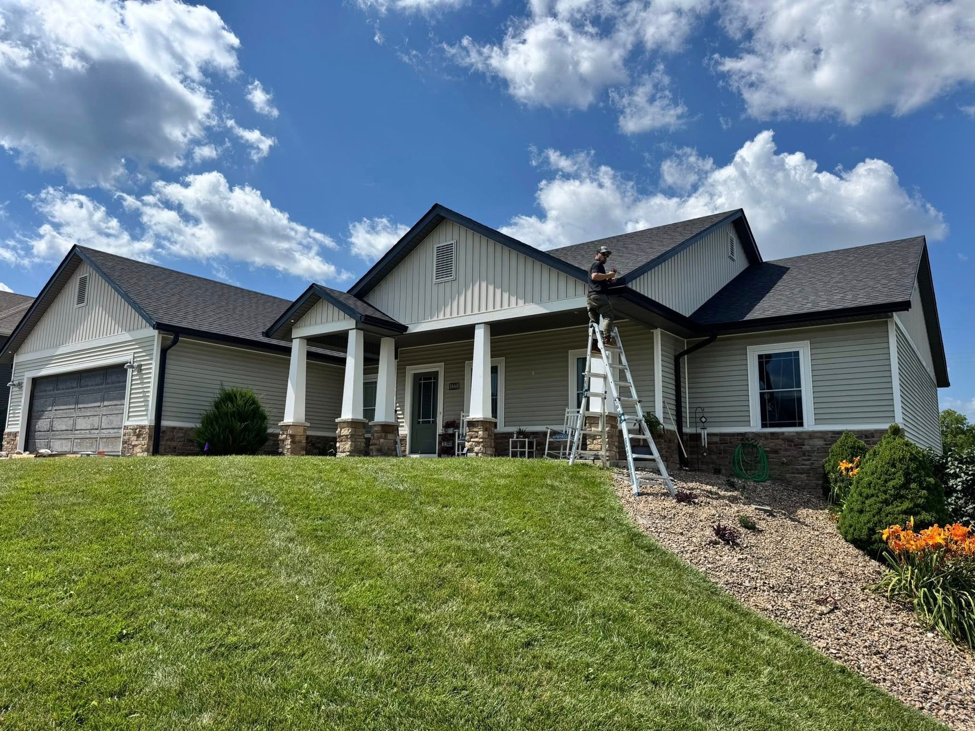 House with tan siding, black roof, and person on ladder cleaning gutters on a sunny day.