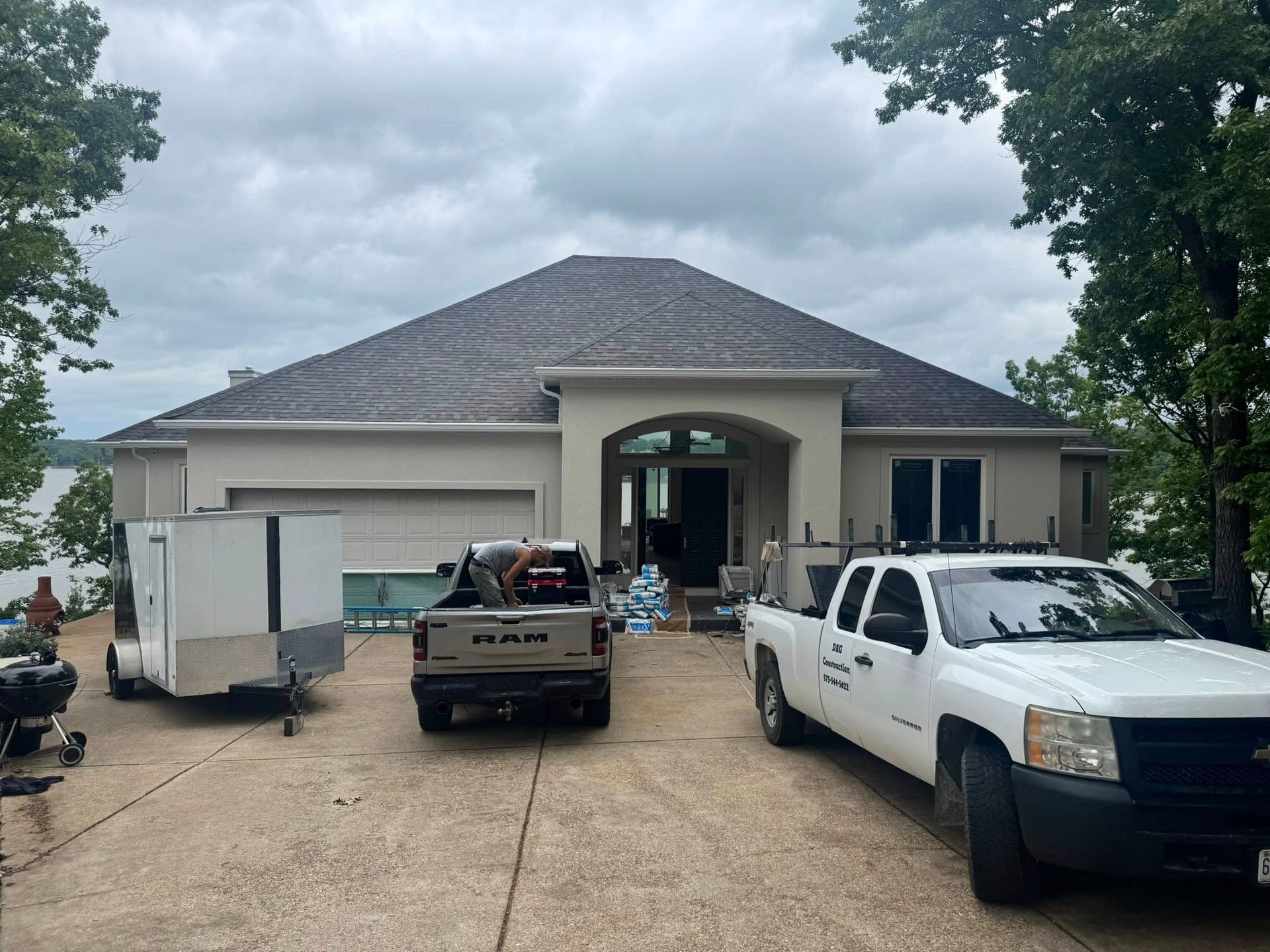 A house with a dark roof and beige walls; work trucks and a trailer are parked in the driveway.