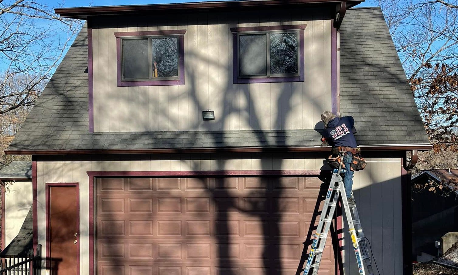 Person on ladder near garage, trimming eaves. Beige building, brown garage door and trim. Sunny day.