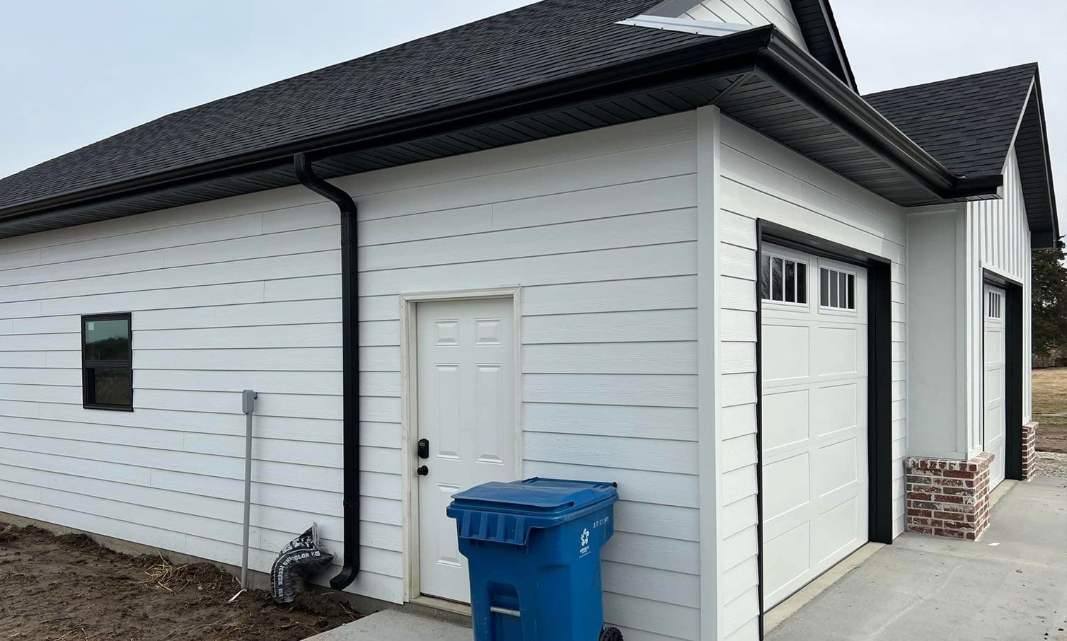 White house with black trim, black roof, and a blue trash bin.