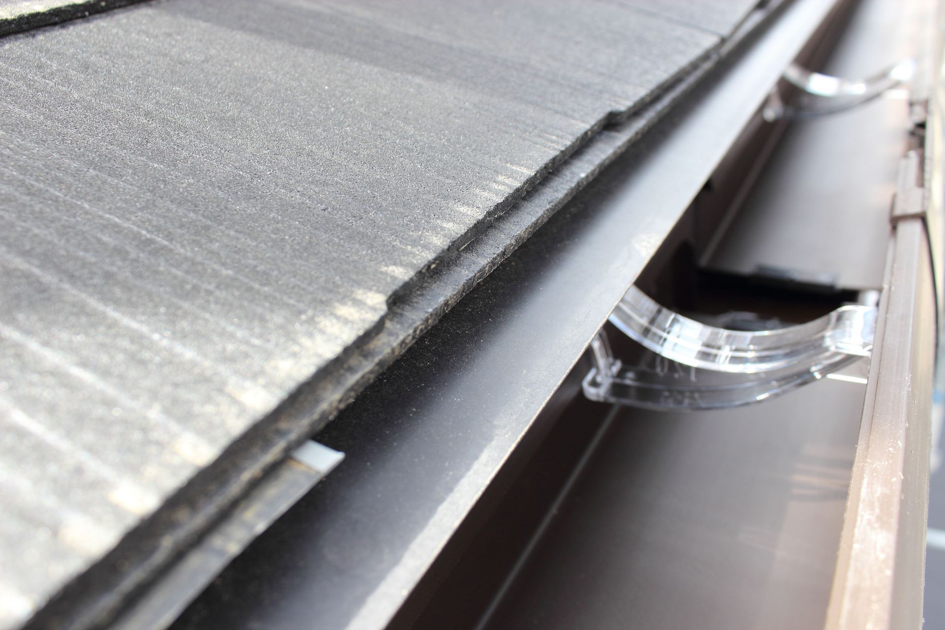 Close-up of a roof edge with shingles and a gutter, showing its brackets and some debris.