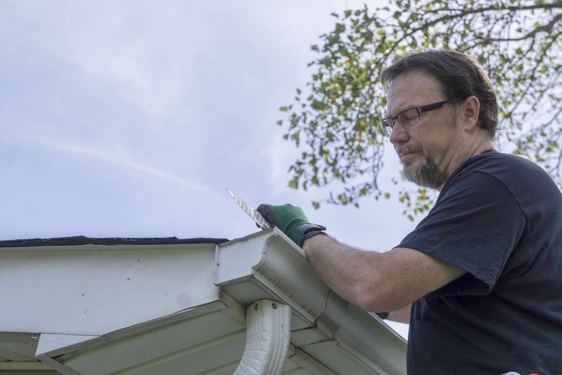 Man wearing glasses and gloves cleaning a white gutter, reaching up outdoors under a cloudy sky.