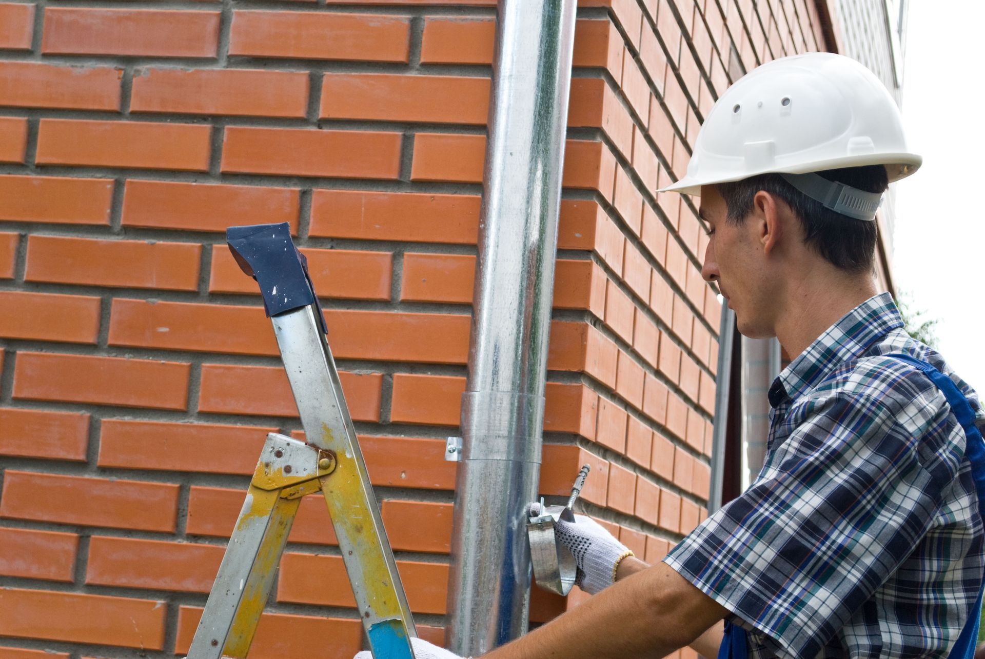 Man in hard hat installing metal pipe on brick wall, using a ladder.