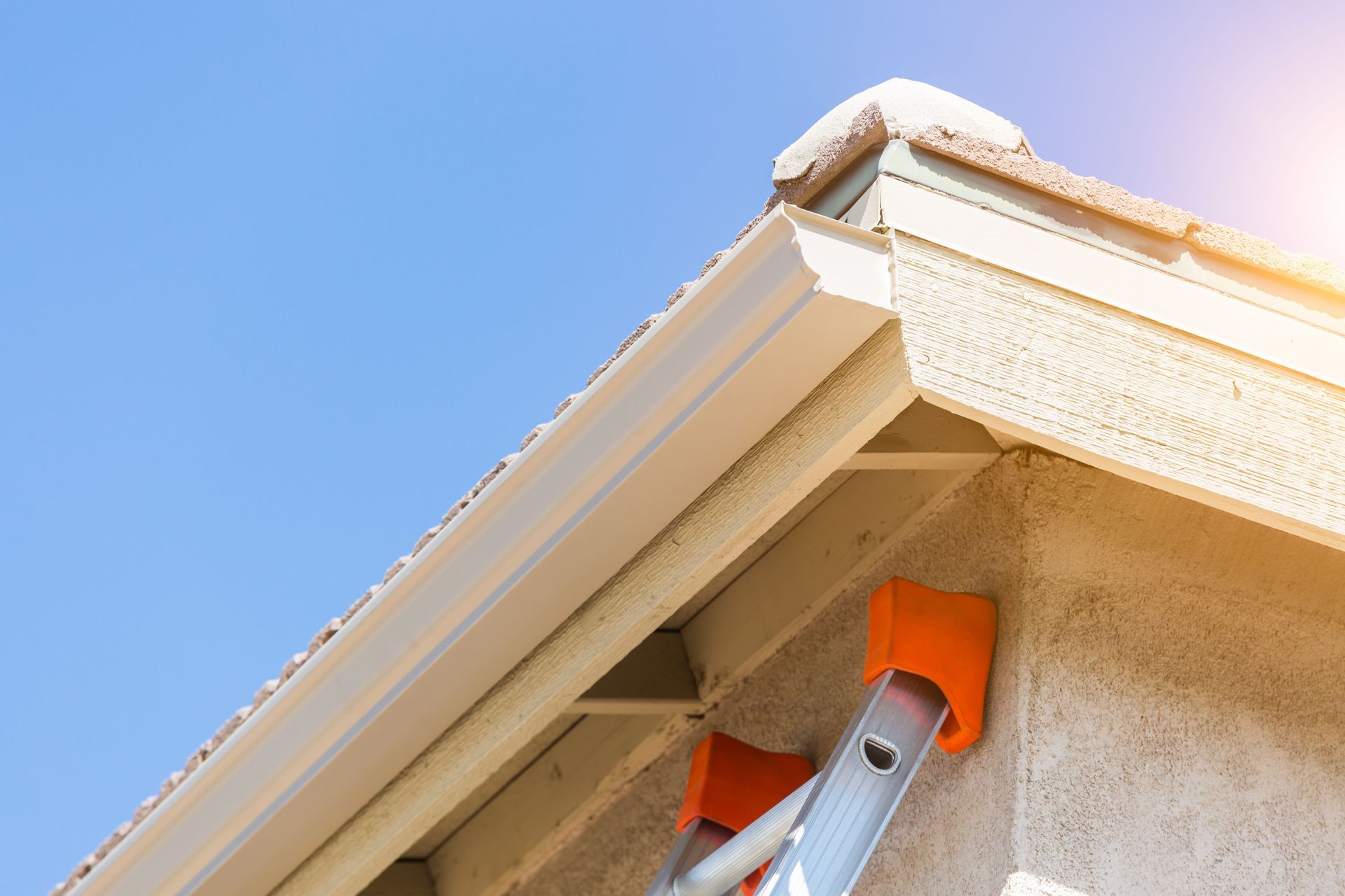 Beige house corner with gutter, ladder, and a clear blue sky.