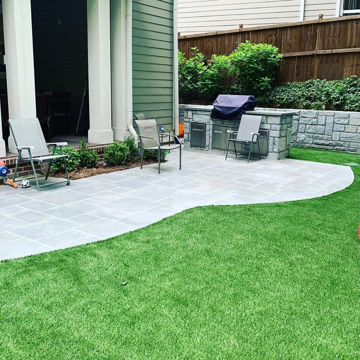 Patio with grill, chairs, and green lawn. Grey pavers, stone wall, and covered area.
